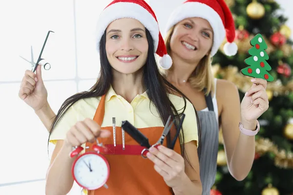 Due donne di parrucchiere in cappelli di Babbo Natale tengono forbici e pettini con sveglia su sfondo di albero Di Capodanno — Foto Stock