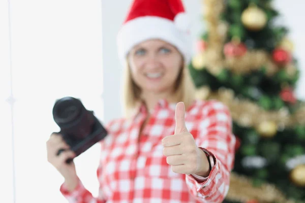 Женщина-фотограф держит фотоаппарат и большой палец вверх жестом на фоне новогодней елки — стоковое фото