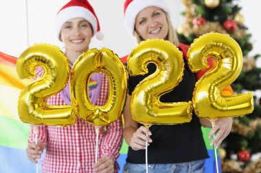 Noel Baba şapkalı iki kadın yeni yıl ağacının arka planında 2022 sayısını tutuyor.