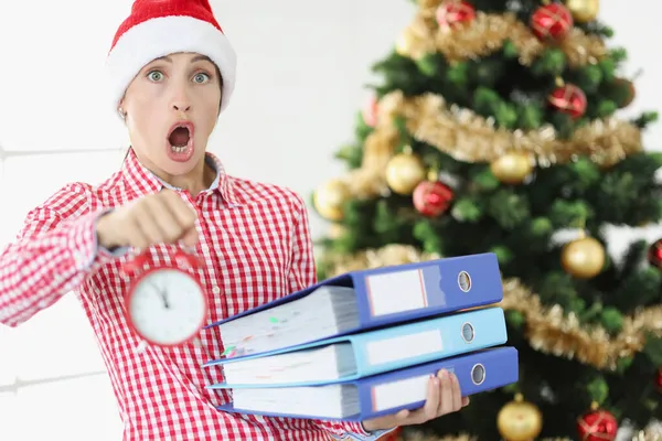 Σοκαρισμένη νεαρή γυναίκα κρατά ξυπνητήρι και στοίβα από φακέλους με έγγραφα στο φόντο του χριστουγεννιάτικου δέντρου — Φωτογραφία Αρχείου