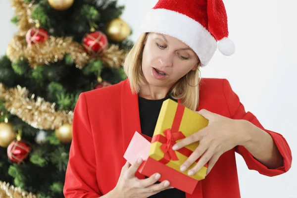 Joven empresaria en chaqueta roja y sombrero de Santa Claus abre caja de regalo contra árbol de Año Nuevo —  Fotos de Stock