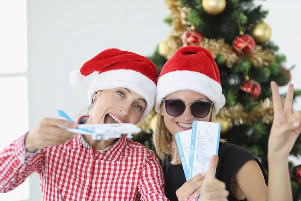 Noel Baba şapkalı iki genç kadının portresi. Elinde uçak ve yılbaşı ağacı biletleri var. — Stok fotoğraf