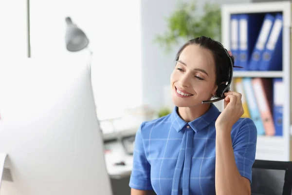 Mujer joven en auriculares con micrófono sentado detrás de la computadora portátil y sonriendo —  Fotos de Stock