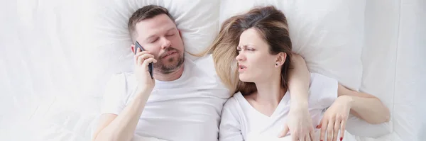 Genç adam karısıyla yatakta uzanıyor ve cep telefonuyla konuşuyor. — Stok fotoğraf