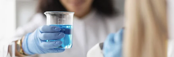 Kobieta chemik trzyma szklaną zlewkę z niebieskim płynem przed mikroskopem w laboratorium — Zdjęcie stockowe