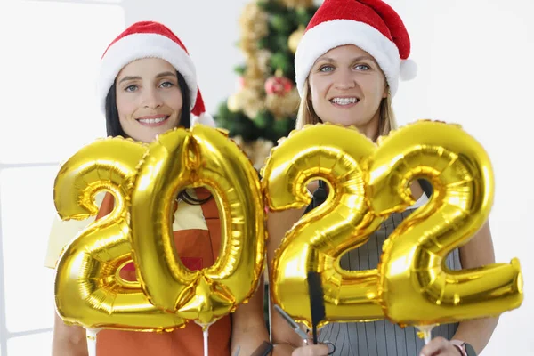 Две улыбающиеся женщины-стилиста держат номера 2022 на фоне новогодней елки. — стоковое фото