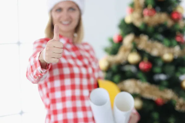 Mujer sonriente arquitecto de diseño sostiene pulgares hacia arriba contra el fondo del árbol de Año Nuevo —  Fotos de Stock