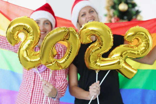 Две улыбающиеся женщины держат флаг ЛГБТ и цифры 2022 на фоне новогодней елки — стоковое фото