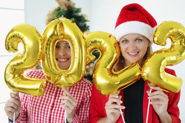 Две улыбающиеся женщины держат номера 2022 на фоне новогодней елки — стоковое фото