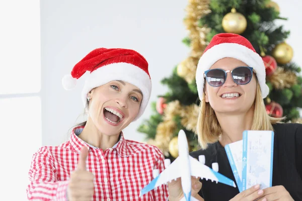 İki neşeli kız arkadaş ellerinde bilet ve yeni yıl ağacının arka planına karşı küçük bir uçak tutuyor. — Stok fotoğraf