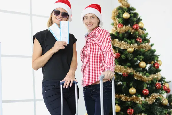 Duas mulheres sorridentes em chapéus de Papai Noel segurar bilhete de avião contra fundo da árvore de Natal — Fotografia de Stock