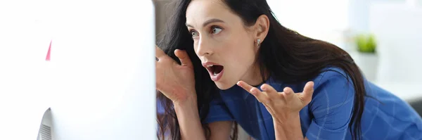 Шокована жінка сидить за столом і дивиться на екран комп'ютера очі, що розширюються в жаху — стокове фото