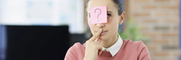 Pensive young woman sitting at table with sticker with question mark on her forehead — Stock Photo, Image