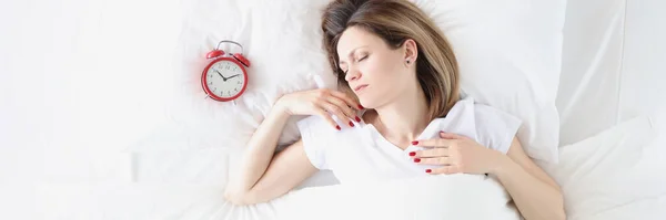 Νεαρή γυναίκα ξαπλωμένη στο κρεβάτι κοντά στο κόκκινο ξυπνητήρι — Φωτογραφία Αρχείου