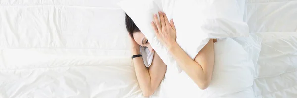 Young woman lying in white bed and covering her ears with pillow top view — Stockfoto