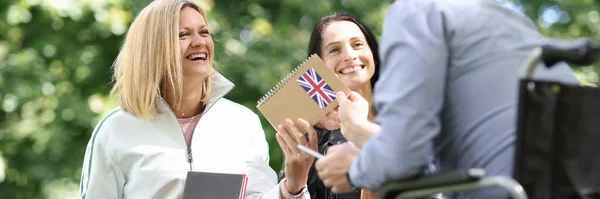 Disabled man in wheelchair giving english textbook to his female friends in park — Stock Photo, Image