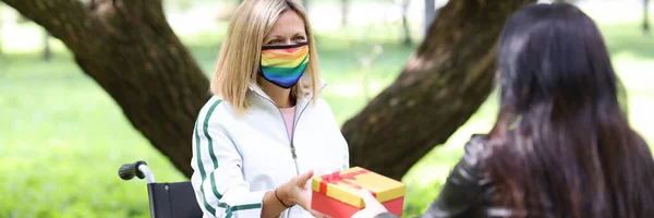 Vrouw geeft geschenk in rode doos aan gehandicapte vriend in beschermende medische masker met lgbt symbolen in het park — Stockfoto