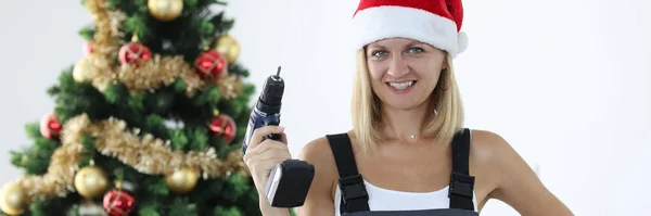 Üniformalı gülümseyen kadın tamirci ve Noel Baba şapkası Noel ağacının yanında elinde matkap tutan. — Stok fotoğraf