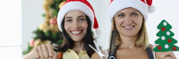 Twee lachende vrouwen kappers in uniformen en kerstman hoeden houden wekker schaar met kam. — Stockfoto