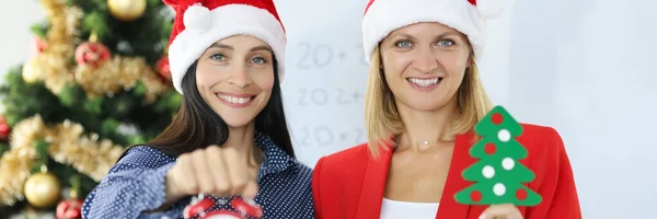 Noel Baba şapkalı iki gülümseyen iş kadını Noel ağacının arka planında çalar saat tutuyor. — Stok fotoğraf