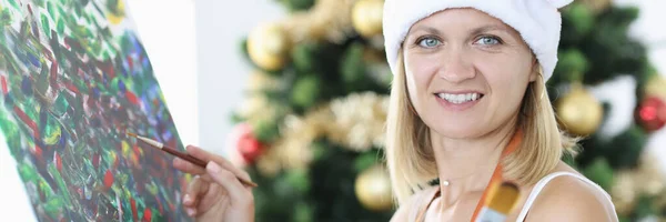 Mujer sonriente artista en sombrero de Santa Claus dibuja imagen sobre el fondo del árbol de Navidad —  Fotos de Stock