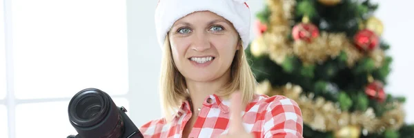 Γυναίκα φωτογράφος στο καπέλο Σάντα Claus στο φόντο του χριστουγεννιάτικου δέντρου κάνει τους αντίχειρες προς τα πάνω χειρονομία — Φωτογραφία Αρχείου