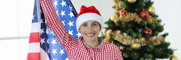 Vreugdevolle vrouw in kerstman hoed houdt vlag van Amerika tegen de achtergrond van Nieuwjaar boom — Stockfoto
