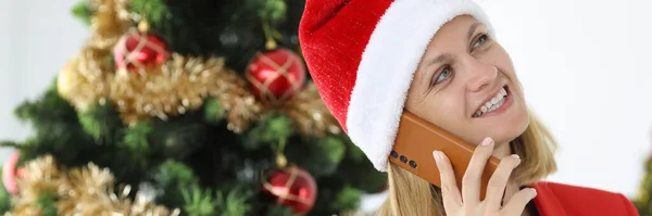 Noel Baba şapkalı kadın telefonda Noel ağacının arka planında konuşuyor. — Stok fotoğraf