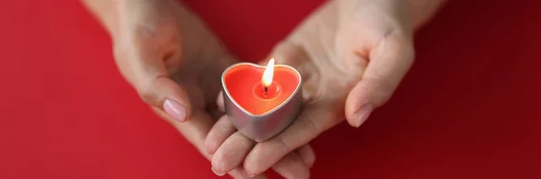 Rött brinnande ljus i form av ett hjärta i kvinnliga händer på röd bakgrund — Stockfoto