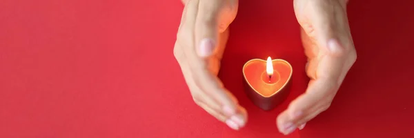 Kvinnlig hand skyddar försiktigt brinnande ljus i form av hjärta på röd bakgrund — Stockfoto