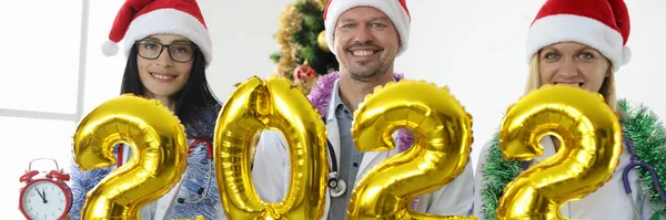 A equipe de doutores em gorros vermelhos mantém o número 2022 contra o contexto da árvore de Ano Novo — Fotografia de Stock
