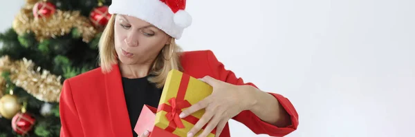 Kırmızı Noel Baba şapkalı genç kadın yılbaşı hediyesini ağacın yanında açıyor ve şaşırıyor. — Stok fotoğraf