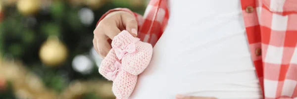 Schwangere hält Baby-rosa Pantoffeln auf Hintergrund des Neujahrsbaums — Stockfoto