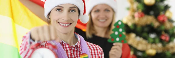 Две улыбающиеся женщины с флагом lgbt держат будильник на фоне новогодней елки — стоковое фото