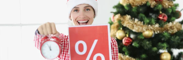 Joven mujer sonriente en sombrero de Santa Claus sostiene reloj despertador y cartel con por ciento en el fondo del árbol de Año Nuevo —  Fotos de Stock