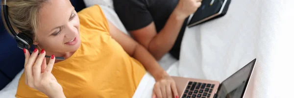 마이크를 가지고 있는 헤드폰을 사용하는 젊은 여자는 침대에 누워 있는 동안 노트북으로 일한다 — 스톡 사진