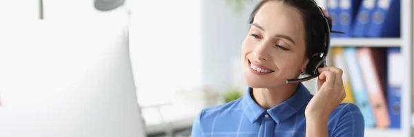 Mujer joven en auriculares con micrófono sentado detrás de la computadora portátil y sonriendo — Foto de Stock