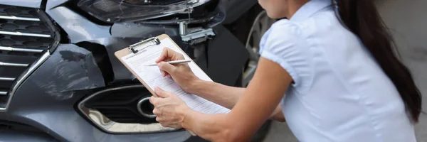 Agen wanita muda yang duduk di dekat mobil rusak dan menulis informasi ke dalam dokumen — Stok Foto