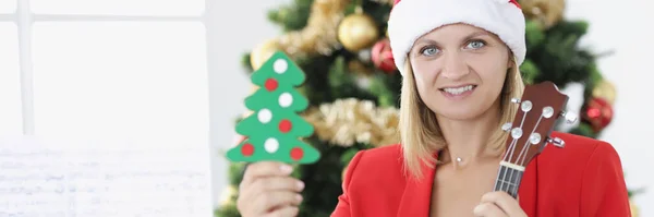 Noel Baba şapkalı kadın müzisyen elinde oyuncak Noel ağacı ve ukulele tutuyor. — Stok fotoğraf