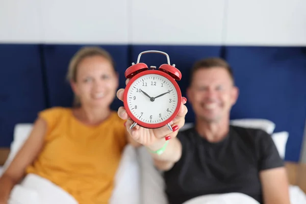 Smiling man and woman lying in bed and holding alarm clock at ten oclock — стоковое фото