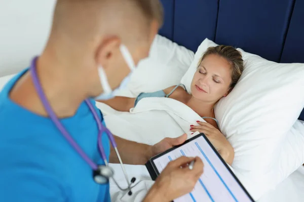 Dokter maakt aantekeningen in patiëntenkaart en onderzoekt patiënt in bed — Stockfoto