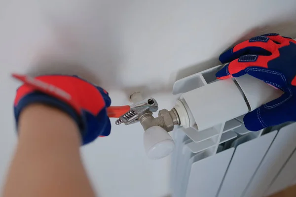 Close-up of plumber in protective gloves installing heating radiator in apartment or house. Builder installs new hot water central heating system using wrench