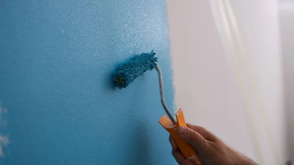 Close-up of male decorator painting wall in blue color. Man painting walls with roller. Repair and renovation of apartments concept