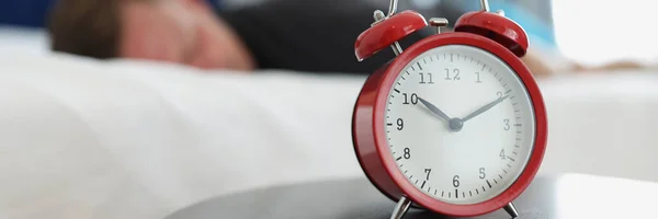 A red alarm clock stands on a round table in the bedroom against the background of a sleeping man, close-up, blurry