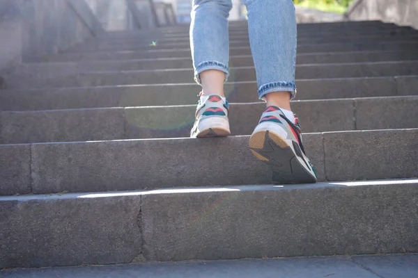 Close-up of woman legs in jeans and sneakers going up steep stairs. Female tourist in casual clothes going upstairs outdoors