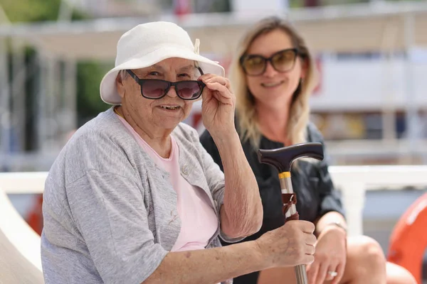 Close-up of old granny with cane sitting on bench with young woman. Grandmother in sunglasses resting outside or waiting public transport. Old age concept