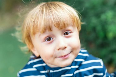 Açık havada küçük sarışın bir çocuğun portresi. Çocuk kameraya bakıyor ve gülümsüyor. Çizgili ceket giyen çocuk güneşli yaz havasında parkta vakit geçiriyor..