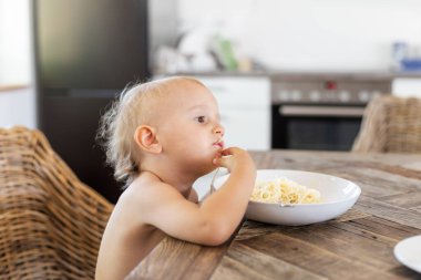 Küçük çocuk mutfakta masada oturuyor ve peynirli spagetti yiyor. Yeni yürümeye başlayan çocuk öğle yemeği yiyor. Çocuk konsepti için sağlıklı yiyecek.