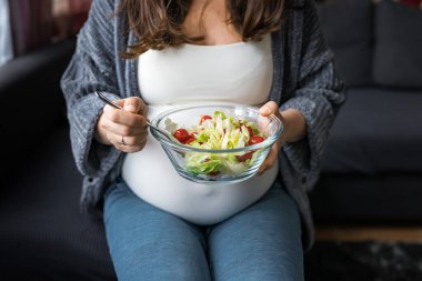 Hamile bir kadın taze sebze salatası yiyor, koltukta oturuyor, sağlıklı hamilelik kavramı.