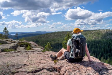 Caucasian outdoor active young adult woman sitting on Rock With outdoor hiking clothes and cap smiling and looking at camera with mountain and lake background in Skuleskogen national park, Sweden.